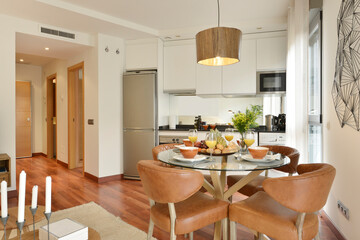 Living room of the vacation rental house with a round glass dining table, brown upholstered chairs and in the background a wall with an open kitchen with reddish wooden floors