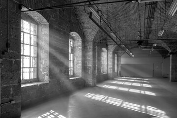 Black and white of long empty room in a penitentiary with limestone walls and arched brick ceilings, steel paned windows with sun streaming in, nobody
