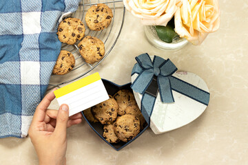 homemade chocolate Chip Cookies on grid and on heart box for valentine's day with napkin and beautiful roses flowers on marble background and woman holding an empty card 