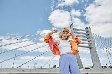 Waist up portrait of free young woman wearing colorful clothes dancing on city bridge and looking...