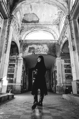 Dark gothic woman with black dress in large ancient abandoned mausoleum in neoclassical style with niches, arches and sunlight (in black and white)