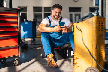 Young male mechanic sitting in workshop and using smart phone. He is smiling.