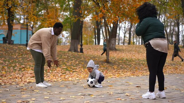Wide shot happy African American man woman and boy playing football on park alley laughing. Smiling father mother and toddler son resting together enjoying autumn leisure