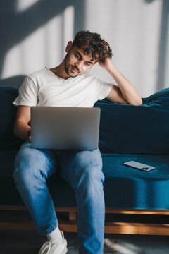 Relaxed young man working on laptop pc computer sitting on blue sofa, spending time in living room at home. nternet communication. Modern communication