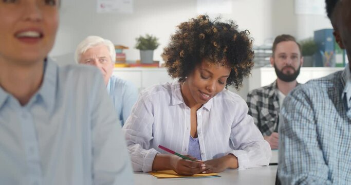 Portrait of african young student writing down notes and raising hand to ask question in classroom