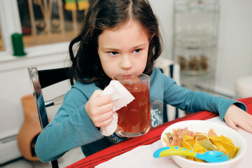 a pretty young girl drinking herbal tea while also eating colorful bow pasta at the dinner table