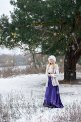 Bright color photo of a beautiful young lady dressed in vintage clothes in a fur hat and vest in a long purple skirt in a winter coniferous forest with pines