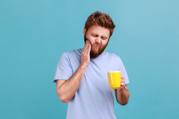 Portrait of bearded man with frowning face standing and touching his cheek because feeling pain on tooth, after drinking cold water. Indoor studio shot isolated on blue background.