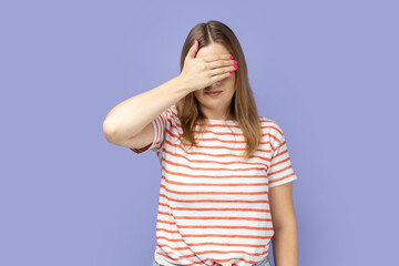 Don't want to look at this. Portrait of blond woman wearing striped T-shirt covering eyes with hand, feeling shamed and scared to watch. Indoor studio shot isolated on purple background.