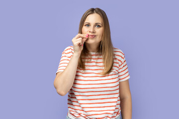 Portrait of beautiful positive blond woman wearing striped T-shirt standing and zipping her mouth, knows secret, does not tell to anyone. Indoor studio shot isolated on purple background.