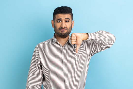 Portrait of young adult businessman criticizing bad quality with thumbs down displeased grimace, showing dislike gesture, wearing striped shirt. Indoor studio shot isolated on blue background.