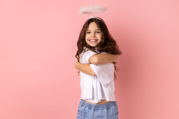 Portrait of smiling little girl wearing white T-shirt and with halo over head embracing herself lovingly and smiling from pleasure. Indoor studio shot isolated on pink background.