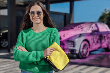Portrait of young female in green sweater with yellow microfiber cloth and car that covered with...