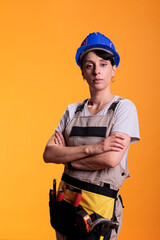 Portrait of female construction worker posing with arms crossed in studio. Professional woman in construction industry looking confident, standing in renovation overalls over yellow background.
