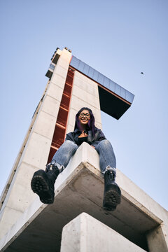 low angle shot of young brunette latina woman wearing big boots blue jeans sitting in the corner with legs in the air smiling at camera next to concrete building