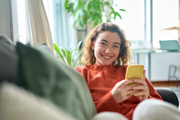 Smiling young pretty woman sitting on couch using apps on cell phone technology, happy lady holding...