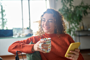 Young happy woman using cell phone drinking coffee relaxing at home. Smiling pretty lady holding...