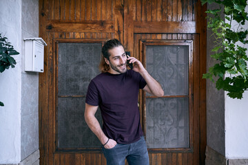 caucasian young man talking on the smartphone of him smiling next to the wooden door and the mailbox with his hand in his pocket