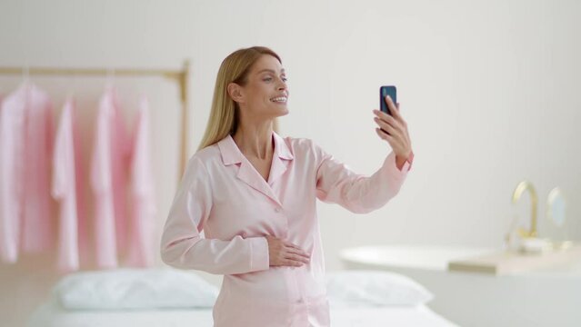 Positive pregnant woman wearing pajamas video chatting with family or friends, standing at home in morning, free space