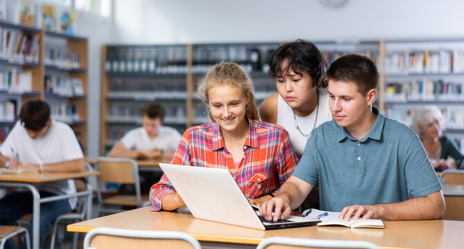 Smiling teenage boy and girls using laptop at college library, watching videos