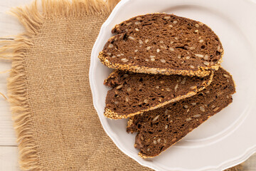 Several pieces of rye multigrain bread with a white plate and jute napkin on a wooden table, macro, top view.