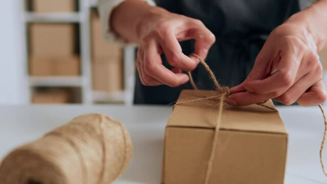 Warehouse worker packs a box for shipment using thread and kraft paper packaging materials, storekeeper works in a warehouse of boxes and cargo packaging, E-commerce inside view
