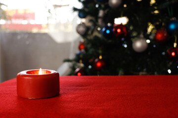 Close-up of burning red candle on a red cloth with defocused christmas tree and window background