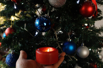 Hand holding a red candle lighting up the Christmas tree decorated with spheres, bows and ribbons