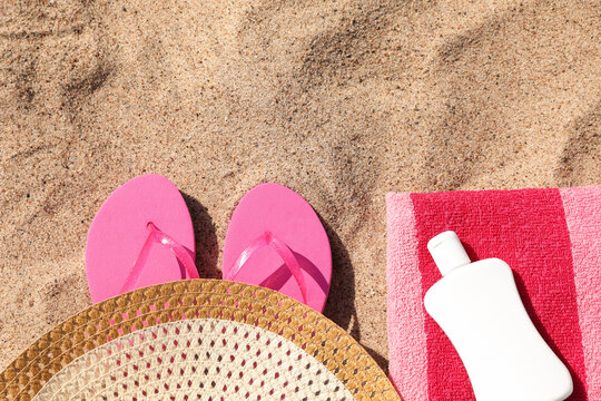 Beach towel with slippers, straw hat and cosmetic product on sand, flat lay. Space for text