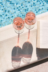 Glasses of tasty rose wine and open book on swimming pool edge, above view