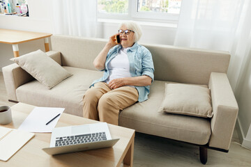 a happy, relaxed elderly woman with short white hair is sitting on a cozy sofa in an apartment and talking on a smartphone stylishly dressed in a light shirt and with glasses on her face