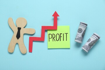 Sticky note with word Profit, rolled banknotes, up arrow and wooden human figure on turquoise...