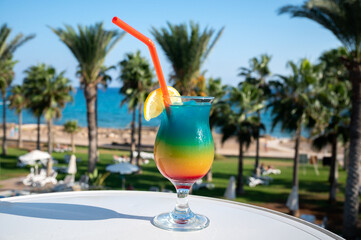 Colourful cold Rainbow Paradise cocktail drink served in glass at pool bar overlooking blue pool, sea and palm trees, relax and holidays at sea