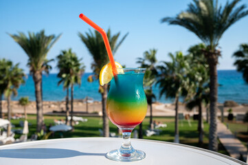 Colourful cold Rainbow Paradise cocktail drink served in glass at pool bar overlooking blue pool, sea and palm trees, relax and holidays at sea
