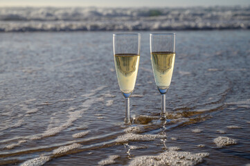 Glasses of cava or champagne sparkling wine on white sandy ocean beach with water waves on sunset in sunlights