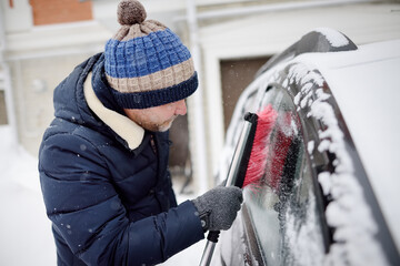 Mature man cleaning car with brush after snowfall. Snowdrift after snowstorm on the hood of automobile. Difficult transport situation on roads in city after snow storm. Winter weather.