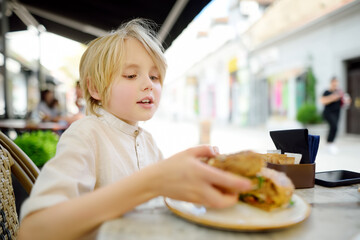 Cute blonde boy eating large bagel with salmon and arugula in outdoor fast food restaurant