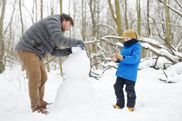 Fototapeta na wymiar Little boy with his father building snowman in snowy park. Active outdoors leisure with family with children in winter. Stroll in a snowy winter park