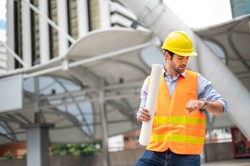 Caucasian man engineers use a smartphone for talking, wearing an orange vest and a big hard hat, and the other hand holding the white floor plan in the site work of the center city.