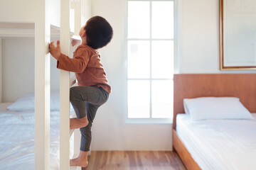 Fototapeta na wymiar Asian little toddler boy is trying to climb up the ladder of bunk bed in the bedroom. Concept of childhood, kid development, physical activity, muscle, playful, learning, life skill for children.