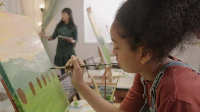 A Black girl concentrates on acrylic color picture painting on canvas with multiracial kids in an art classroom, creative learning with talents and skills in the elementary school studio education.