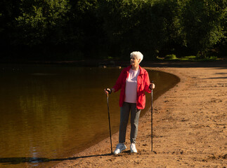 Grey-haired woman walking with tracking sticks on the beach near lake