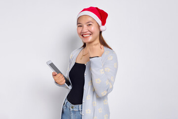 Cheerful young Asian woman in a hat Christmas holding a digital tablet, showing victory isolated over white background. Indonesian people celebrate Christmas concept