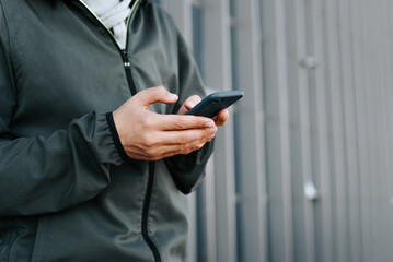Unrecognizable man in jacket using phone while standing against background geometric wall, close-up. Side view of male hands holding smartphone outdoors