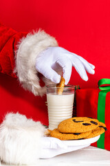 Obraz na płótnie Canvas Hand of Santa Claus with traditional Christmas cookie snack with milk glass. Santa Dunking Cookie, on bright festive red and white wooden background. Merry Christmas and Happy New Year greeting card