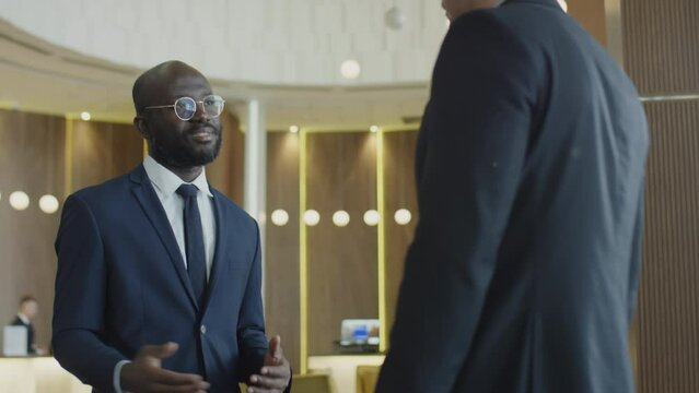 Medium shot of African American businessman greeting Caucasian colleague in hotel lobby with handshake and speaking with him