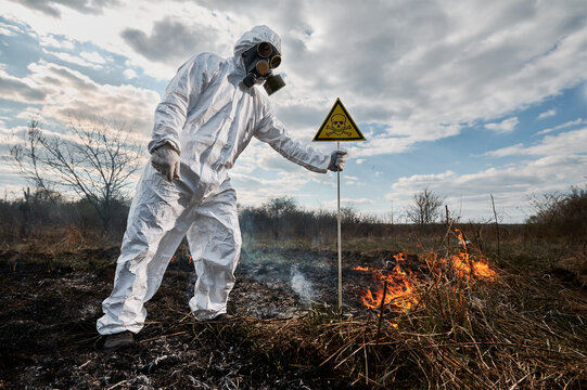 Fireman ecologist fighting fire in field. Man in protective radiation suit and gas mask near burning grass with smoke, holding triangle with skull and crossbones warning sign. Natural disaster concept