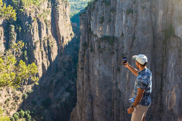 Male in checkered blue shirt and a cap takes a photo on a smartphone of a cliff of a deep canyon, a...