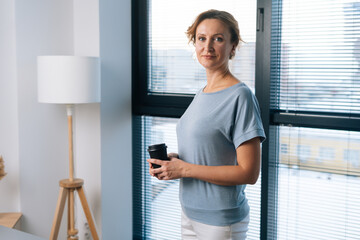 Portrait of elegant business woman holding coffee cup and looking at camera standing on background of large window at office with modern light interior. Confident blonde businesswoman posing indoors.