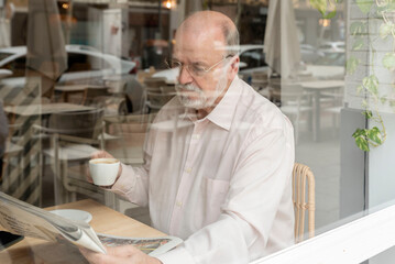 Fototapeta na wymiar an older man in glasses sitting inside a coffee shop behind a window reading a newspaper holding a cup of coffee